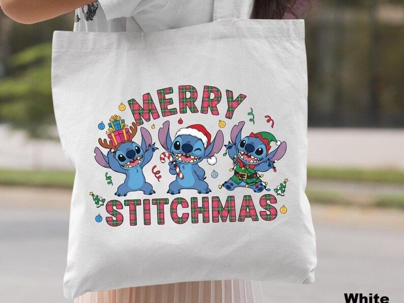 May include: White tote bag with a festive design featuring three blue cartoon characters dressed for Christmas. The words "MERRY STITCHMAS" are displayed above the characters in a plaid font. The bag is held by a person.