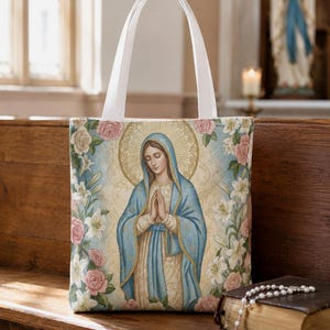 May include: A rectangular tote bag with white fabric handles featuring a central illustration of the Virgin Mary with hands clasped in prayer, surrounded by a decorative border of pink roses and white lilies against a pale gold background.