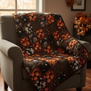 May include: A Halloween-themed quilt draped over a gray armchair. The quilt features a patchwork design with orange pumpkins, white ghosts, and black skulls on a black and green background. The quilt is a cozy home decor item.