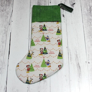 May include: A Christmas stocking with a green cuff and a loop for hanging. The main body of the stocking is white with a pattern of Mickey Mouse, Donald Duck, Christmas trees, and the words "Merry Christmas".