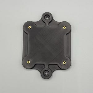 May include: A black, square-shaped component with rounded corners and four brass-colored fasteners. The component has a textured surface and two circular openings on opposite sides. The overall design is symmetrical and industrial.