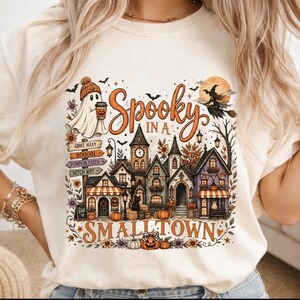 May include: A cream-colored t-shirt featuring a graphic of a small town with a clock tower, a ghost holding a coffee cup, and a witch on a broomstick. The text reads Spooky in a Small Town, with signs for Ghost Alley, Pumpkin Patch, and Witch Way.
