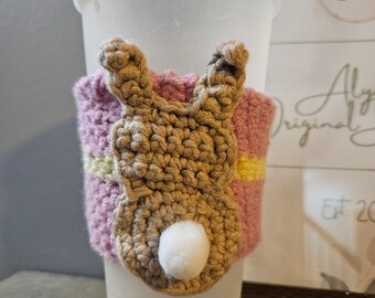 Op de afbeelding: Een roze en gele gestreepte gehaakte bekerwarmer met een bruin konijn ontwerp. Het konijn heeft een witte pompon staart.