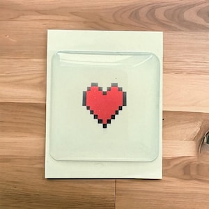 Pode incluir: Uma bandeja quadrada branca com um inserto de vidro transparente. O inserto de vidro tem um design de coração pixelado vermelho.
