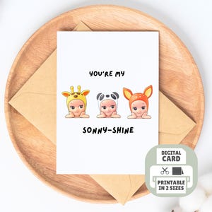 Puede incluir: Una tarjeta de felicitación blanca con el texto "YOU'RE MY SONNY-SHINE" e ilustraciones de tres bebés con disfraces de animales: una jirafa, un panda y un ciervo. La tarjeta está sobre una bandeja de madera con un sobre marrón.
