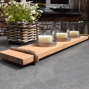 Könnte beinhalten: Ein Holztablett mit einem Seilgriff und vier Glaskerzenhaltern mit weißen Kerzen. Das Tablett befindet sich auf einer grauen Oberfläche.