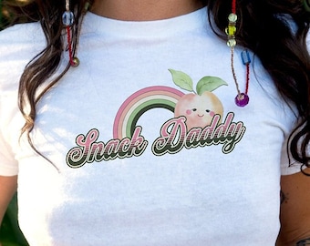 May include: White t-shirt featuring a peach graphic and the text "Snack Daddy" in a retro font.