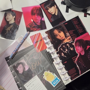 May include: A collection of K-pop merchandise, including photo cards, a planner, and stickers. The photo cards feature images of male performers. The planner is open to a page with a photo and a to-do list. A pen and other stationery items are also visible.