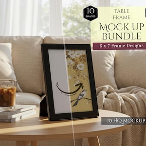 May include: A black 5x7 inch table frame mockup is displayed on a wooden table. The frame holds a print with a bird and floral design. The scene includes a candle, a glass of iced coffee, and a stack of books, suggesting a cozy interior setting. The text on the image says "Table Frame Mock Up Bundle".