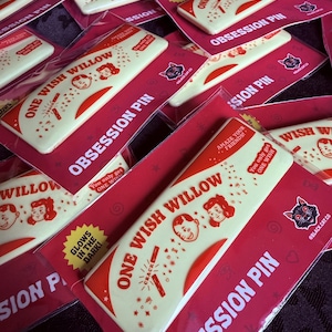 May include: A rectangular off-white pin featuring red text that reads ONE WISH WILLOW and OBSESSION PIN. The design includes illustrations of two faces, a cracked stick, and stars. A yellow starburst reads GLOWS IN THE DARK.