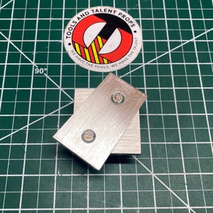 Puede incluir: Primer plano de dos bloques de metal cepillado, uno rectangular y otro cuadrado, con dos tornillos visibles en el bloque rectangular. Una pegatina con el texto "TOOLS AND TALENT PROPS" está en el fondo. El fondo es una esterilla de corte verde.