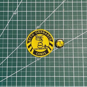 Puede incluir: Dos parches bordados, uno grande y otro pequeño, con fondo amarillo y contornos negros. El parche grande presenta el texto "ECTO ORDNANCE CORPS" y una imagen de dibujos animados de un vehículo de cazafantasmas con el texto "DON'T SLIME ON ME". El parche pequeño presenta una serpiente con el texto "ECTO CORPS".