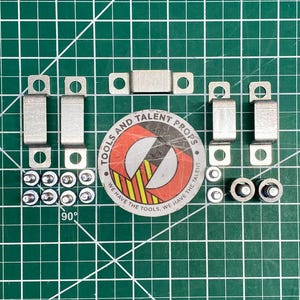 Peut inclure: Divers composants de quincaillerie métallique, comprenant des supports, des vis et des fixations, disposés sur une surface quadrillée verte. Un autocollant circulaire avec le texte "TOOLS AND TALENT PROPS" est également présent. Les composants sont de couleur argentée.