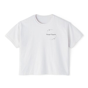 May include: A white short-sleeved t-shirt featuring a small graphic on the upper left chest. The graphic consists of the text "Midnight Margaritas" in a black cursive script, surrounded by a circular arrangement of small black stars and dots.