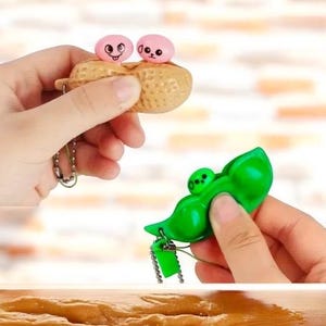 May include: Two keychain toys, one shaped like a peanut with two pink faces and the other shaped like a green pea pod with two green faces.