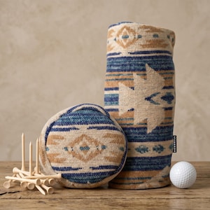 May include: A cylindrical golf club head cover and a round golf club head cover featuring a woven pattern in shades of blue, tan, and cream. A small black tag with the text "HURNHILL" is attached. A pile of wooden golf tees and a white golf ball.