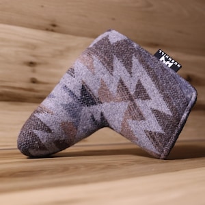 Op de afbeelding: Een golfclub putterhoes met een geometrisch patroon in tinten grijs, bruin en blauw. De hoes heeft een rechthoekige vorm met een afgeronde punt en een klein label met tekst. De hoes is gemaakt van een textuurstof.