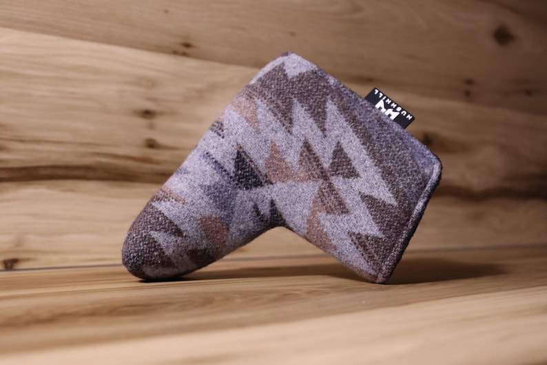 Op de afbeelding: Een golfclub putterhoes met een geometrisch patroon in tinten grijs, bruin en blauw. De hoes heeft een rechthoekige vorm met een afgeronde punt en een klein label met tekst. De hoes is gemaakt van een textuurstof.