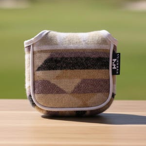 May include: A golf club head cover with a geometric pattern in shades of brown, tan, and black. The cover has a white trim and a small black tag that reads "HUSHHILL". The cover is sitting on a wooden surface.