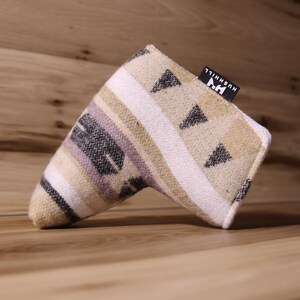 Op de afbeelding: Een golf putterhoes met een geometrisch patroon in beige, wit, zwart en lavendel. De hoes heeft een rechthoekige vorm met een gebogen uiteinde en een klein zwart label met witte tekst "HUSHHILL".