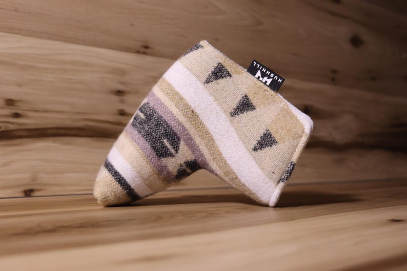 Op de afbeelding: Een golf putterhoes met een geometrisch patroon in beige, wit, zwart en lavendel. De hoes heeft een rechthoekige vorm met een gebogen uiteinde en een klein zwart label met witte tekst "HUSHHILL".