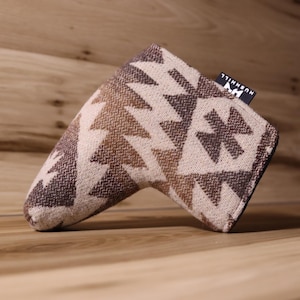 Op de afbeelding: Een golfclub putterhoes met een geometrisch patroon in bruin- en beigetinten. De hoes heeft een rechthoekige vorm met een afgerond uiteinde en een klein label met de tekst "HUSHHILL". De hoes is gemaakt van een geweven stof.