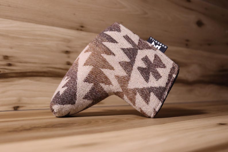 Op de afbeelding: Een golfclub putterhoes met een geometrisch patroon in bruin- en beigetinten. De hoes heeft een rechthoekige vorm met een afgerond uiteinde en een klein label met de tekst "HUSHHILL". De hoes is gemaakt van een geweven stof.