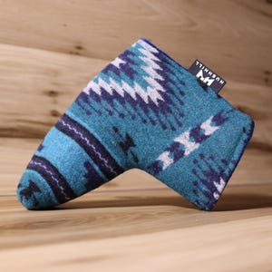 Op de afbeelding: Een teal en marineblauwe golf putterhoes met een geometrisch patroon. De hoes heeft een rechthoekige vorm met een gebogen uiteinde en een klein zwart label met witte tekst "HUSHHILL". De hoes is gemaakt van een zacht, getextureerd materiaal.