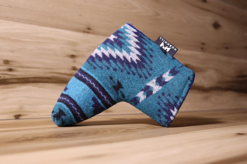 Op de afbeelding: Een teal en marineblauwe golf putterhoes met een geometrisch patroon. De hoes heeft een rechthoekige vorm met een gebogen uiteinde en een klein zwart label met witte tekst "HUSHHILL". De hoes is gemaakt van een zacht, getextureerd materiaal.