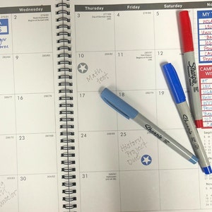 May include: A monthly calendar with a list of college names and a note that says "My Top 8" and "Campus Visit Wishlist". The calendar has a few handwritten notes, including "Math Test" and "History Project Due". There are also two blue highlighters and one red highlighter on the page.
