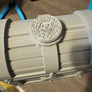 May include: A gray, 3D printed treasure chest with a skull and crossbones design on the lid. The chest has a hinged lid and a decorative metal clasp.