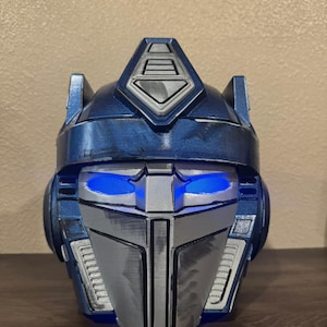 May include: A blue and silver Optimus Prime helmet with glowing blue eyes. The helmet is a detailed replica, featuring intricate paneling and a metallic finish. The design is inspired by the Transformers franchise.