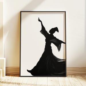 Peut inclure: Impression artistique encadrée en noir et blanc représentant une silhouette d'une personne en robe fluide, les bras levés. L'œuvre est exposée sur un sol en bois, adossée à un mur blanc. L'image est dans un cadre noir.