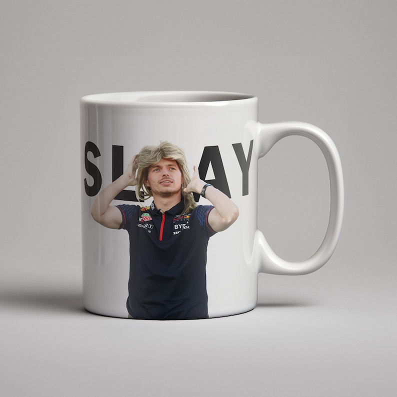 May include: White ceramic mug with a black and white image of a person with blonde hair wearing a black shirt with red and white accents. The text "SLAY" is printed on the mug.