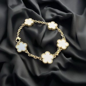 May include: Gold-toned bracelet featuring five white clover-shaped charms. The charms are set in a gold frame and connected by a delicate gold chain. The bracelet is displayed on a black satin background.