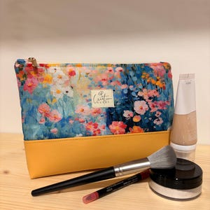 Peut inclure: Une trousse rectangulaire zippée avec un imprimé floral coloré sur la partie supérieure et une base jaune moutarde unie. Une petite étiquette en tissu avec le texte Creation Cathy est centrée. Un pinceau, un crayon rouge et un pot sont présents.