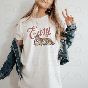 May include: White t-shirt with the word "Easy" in red script and a tiger illustration. The model is wearing a denim jacket.