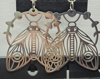 May include: Silver earrings featuring a detailed design of a moth with a circle of thorns around it.