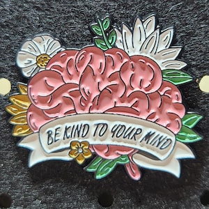 Puede incluir: Pin de esmalte rosa con un cerebro estilizado rodeado de flores y una pancarta que dice "Sé amable con tu mente".