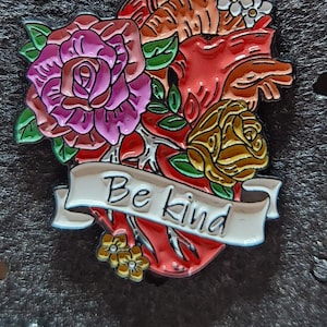 Puede incluir: Un corazón anatómico rojo con una pancarta que dice "Be Kind". El corazón está rodeado de flores, incluyendo una rosa rosa, una rosa amarilla y flores blancas.