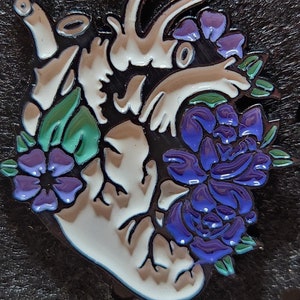 Puede incluir: Un corazón anatómico blanco con un diseño detallado, rodeado de flores moradas y verdes. El corazón está contorneado en negro y tiene un acabado brillante.