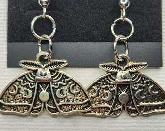 May include: A pair of silver earrings featuring a detailed moth design with crescent moon accents. The earrings have a simple hook closure.