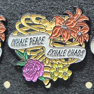 Puede incluir: Un pin de esmalte en forma de corazón con un diseño de caja torácica de esqueleto, flores y el texto "INHALE PEACE EXHALE CHAOS".