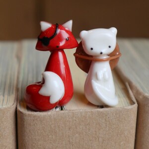 May include: Two small glass animal figurines, a red fox with a black eye patch and a white fox with a brown collar, are perched on the edge of a stack of brown books.