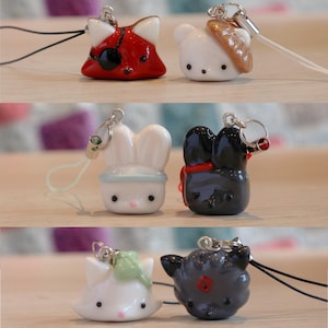 May include: A collection of ceramic animal charms, including a red cat with an eye patch, a white bear with a pastry, a white bunny with a chef's hat, a black figure with a red scarf, a white cat with a green bow, and a gray figure with a red letter G.