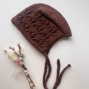 May include: A dark brown knitted bonnet featuring a textured lace pattern and a folded brim. The bonnet includes two long, matching knitted ties attached to the base. Beside the bonnet lies a small decorative bundle of dried pussy willow branches.