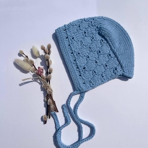 May include: A light blue knitted bonnet featuring a textured diamond pattern and two long chin ties, accompanied by a small bundle of dried pussy willow branches and tiny pink flowers tied together with a piece of natural twine.