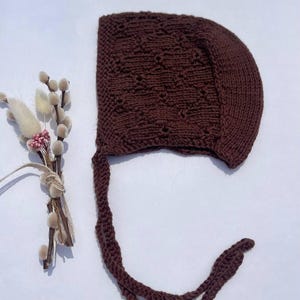 May include: A dark brown knitted bonnet featuring a textured diamond pattern and a smooth border. The bonnet includes two long, matching knitted ties attached at the base for securing the headpiece. The design is uniform in color and material.