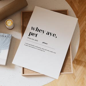 Puede incluir: Una impresión blanca con la frase "whey aye, pet" en negro, junto con su pronunciación y definición. También se ven una taza de café y un libro. La impresión está en un marco de madera.