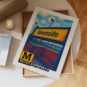 Op de afbeelding: Een ingelijste kunstprint met een kleurrijke illustratie van het metrostation Simonside. Het kunstwerk toont het bord van het station in geel en blauw, met het woord "Simonside" prominent weergegeven. De print bevat ook het Metro-logo.